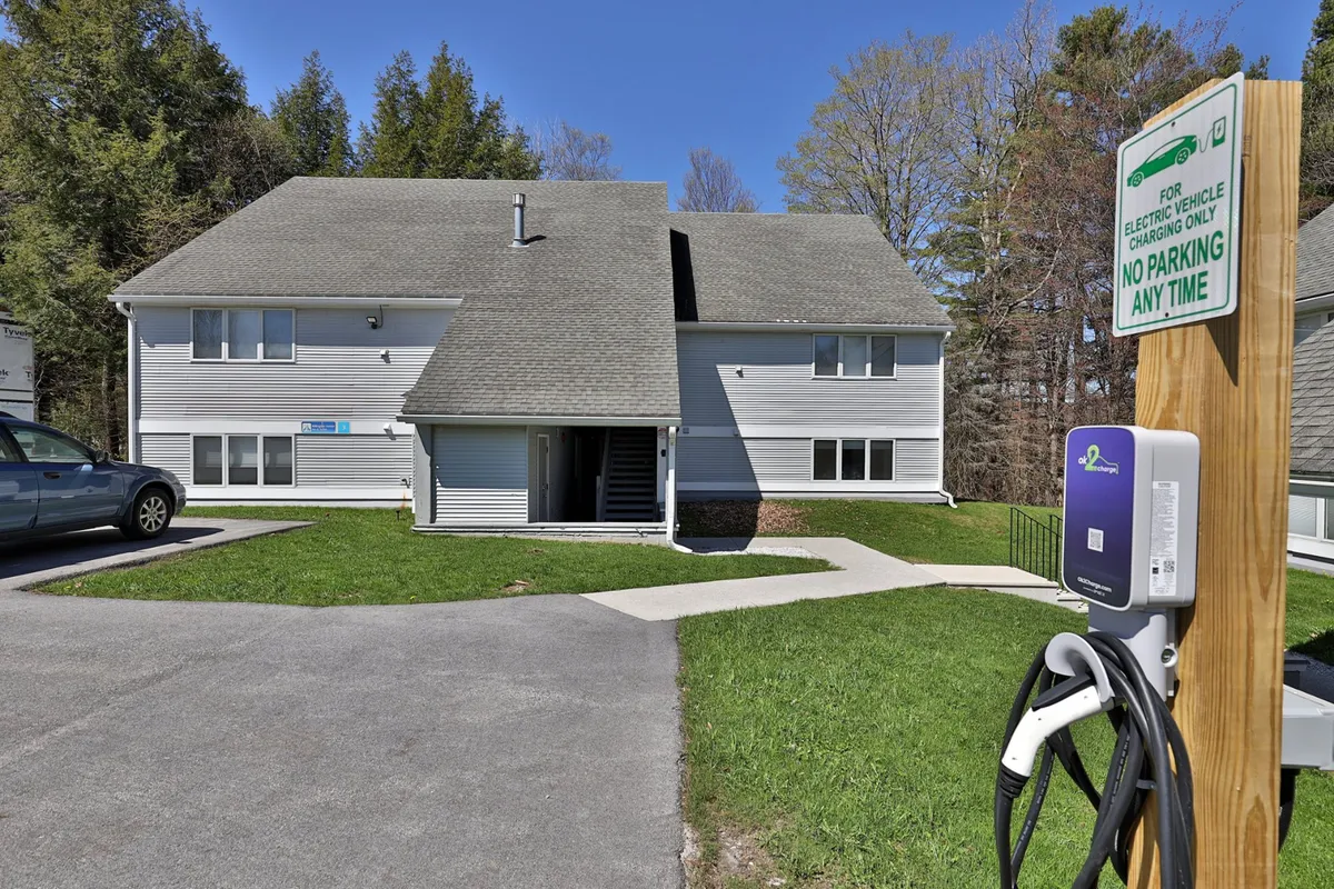 The exterior of the building is shown, highlighting the on-site electric vehicle charging station and a tidy green lawn. The presence of an EV charger aligns with the property's eco-friendly, modern conveniences available to guests.