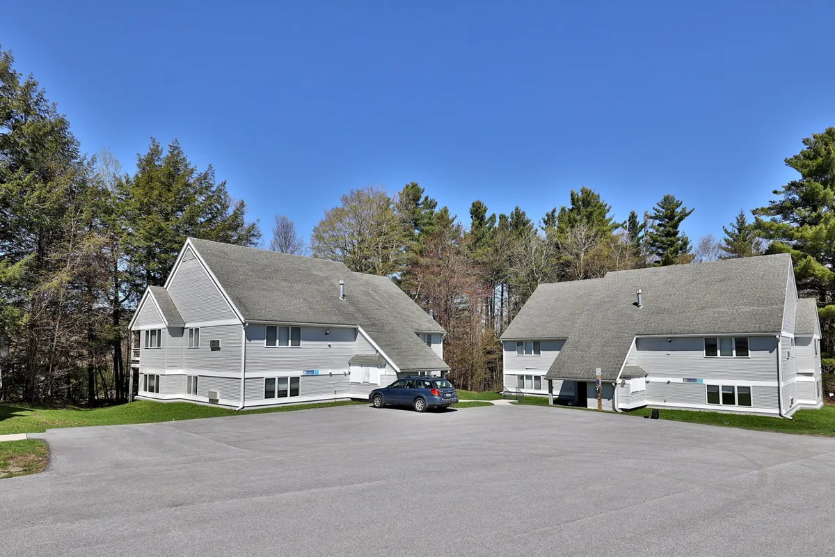 An exterior shot of two residential buildings surrounded by trees, with a visible parking area in the foreground. The natural scenery suggests a peaceful retreat, while the buildings show the type of accommodations guests can expect.