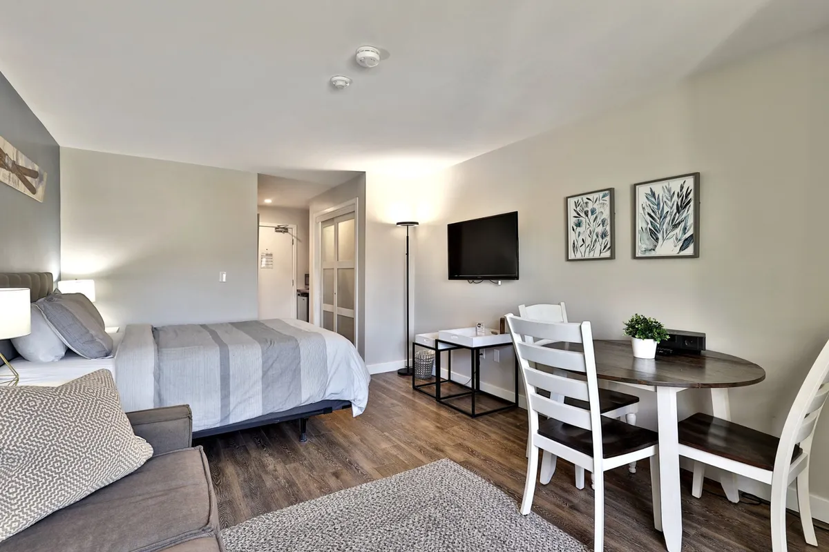 The image showcases a cozy studio interior with a plush queen bed dressed in gray bedding, a comfy sofa adorned with patterned cushions, a small dining table with chairs perfect for intimate meals, a wall-mounted TV for movie nights, and eye-catching wall art that adds warmth to the space. The setting is inviting and well-lit, offering both comfort and functionality for guests.
