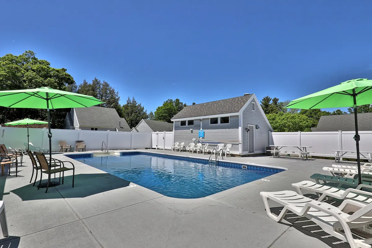A relaxing poolside setting with a clear blue pool, green umbrellas, lounge chairs, a white fence, and leafy trees providing a lush sense of privacy and comfort. The setup invites sunbathing and socializing, with the vibe of a perfect summer afternoon spent outdoors.