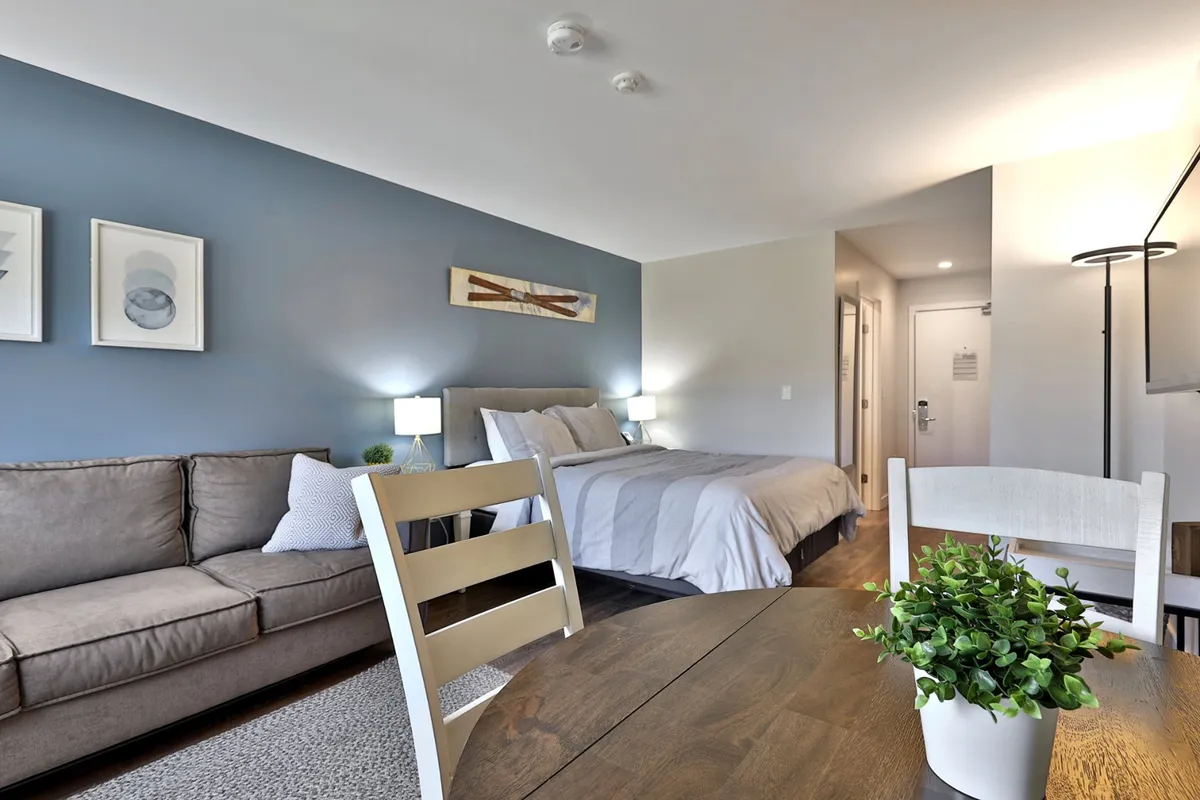 A welcoming interior scene displaying a comfortable bed, a sofa adjacent to a dedicated table that can be used for dining or remote work, and decorative artwork adorning the walls. Soft natural light fills the space, creating a warm and productive atmosphere ideal for both relaxation and focused work sessions.