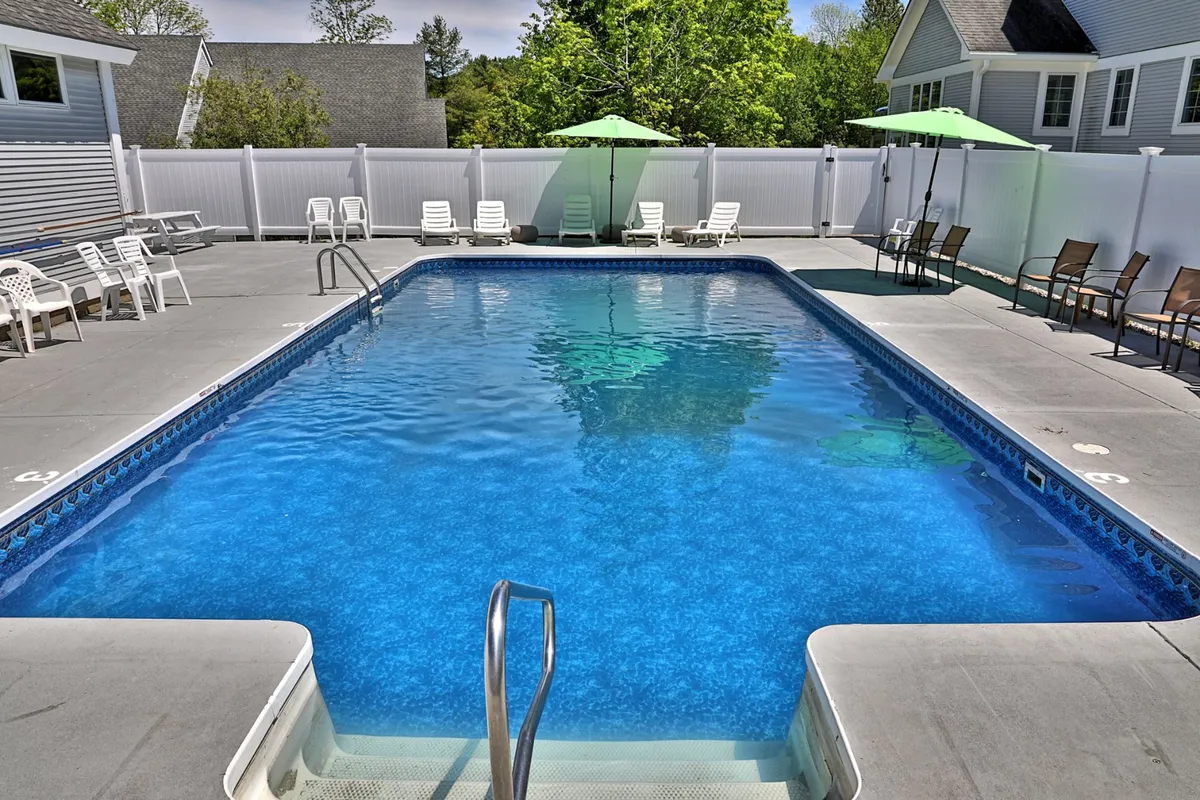 A sunlit outdoor swimming pool is surrounded by lounge chairs and shaded by green umbrellas. The setting is inviting, ideal for relaxation, and conveys the promise of summertime leisure that the heated saltwater pool brings guests.
