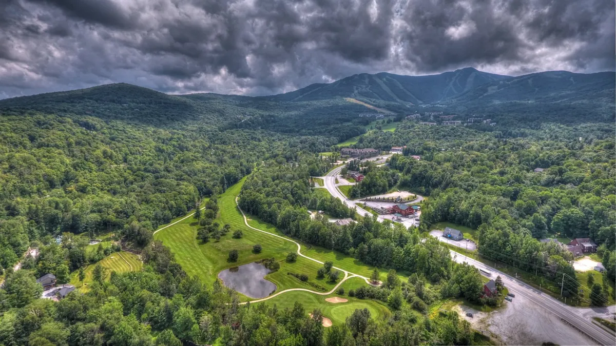 A wide view of mountains, a golf course, a pond, and tree-lined roadways. The landscape is open and scenic, conveying a sense of adventure and proximity to outdoor recreation.